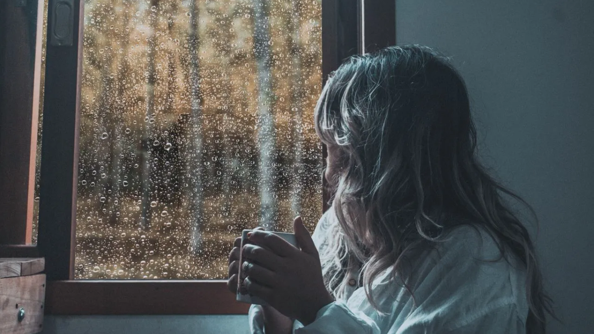 A woman holding a mug and looking out of a rain-streaked window.