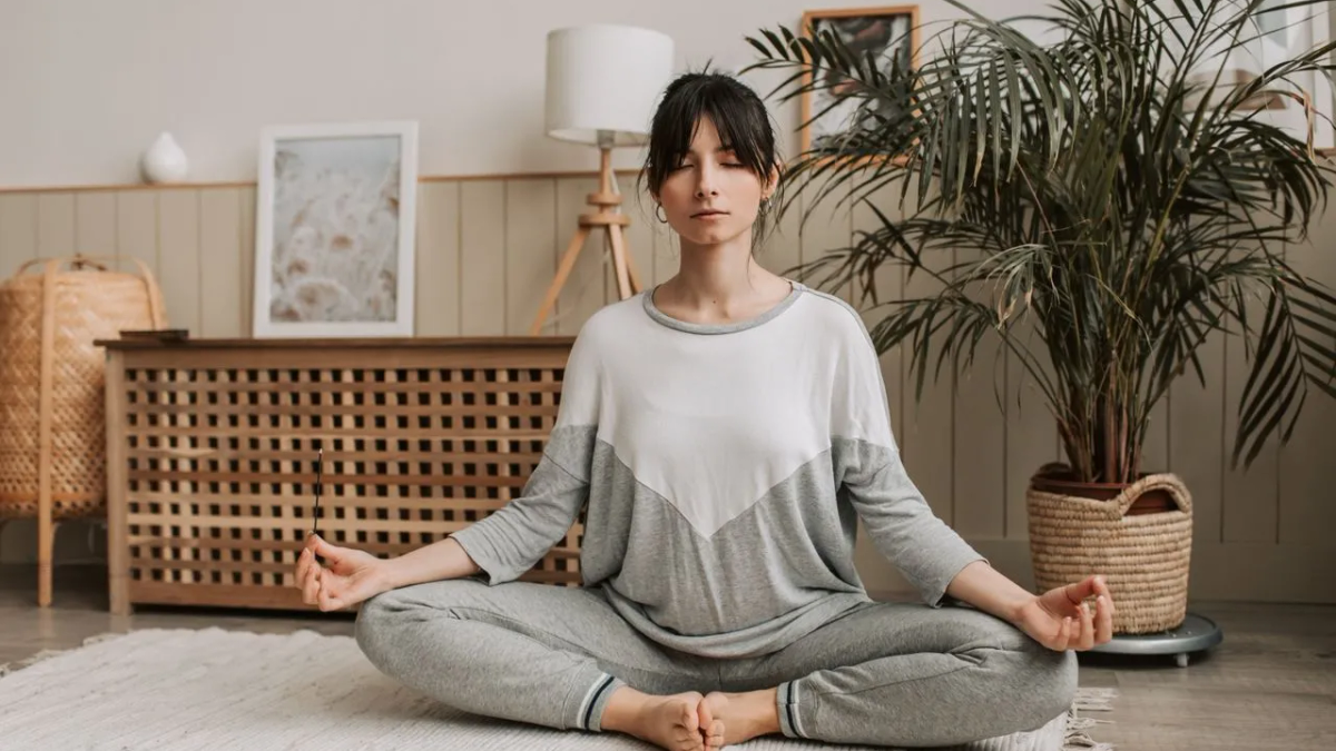 A woman sitting cross-legged on a rug in a calm living room with her eyes closed.