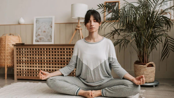 A woman sitting cross-legged on a rug in a calm living room with her eyes closed.