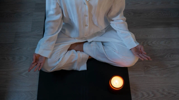 A person in white linen clothing sitting cross-legged in candlelight, hands resting open in a calm meditation pose