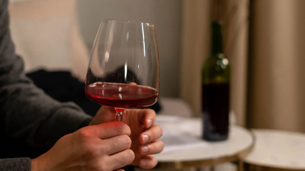 A hand holding a glass of red wine, with the bottle on a table behind.