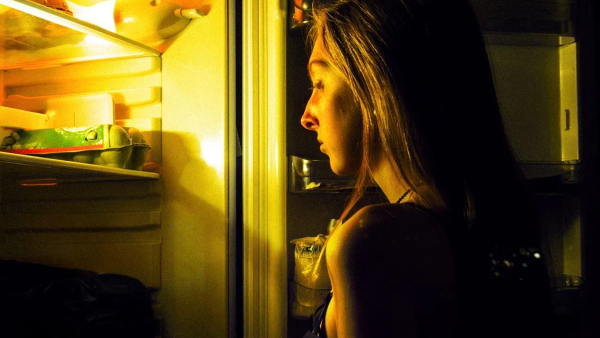 A woman standing at an open fridge at night, lit by the light inside it.